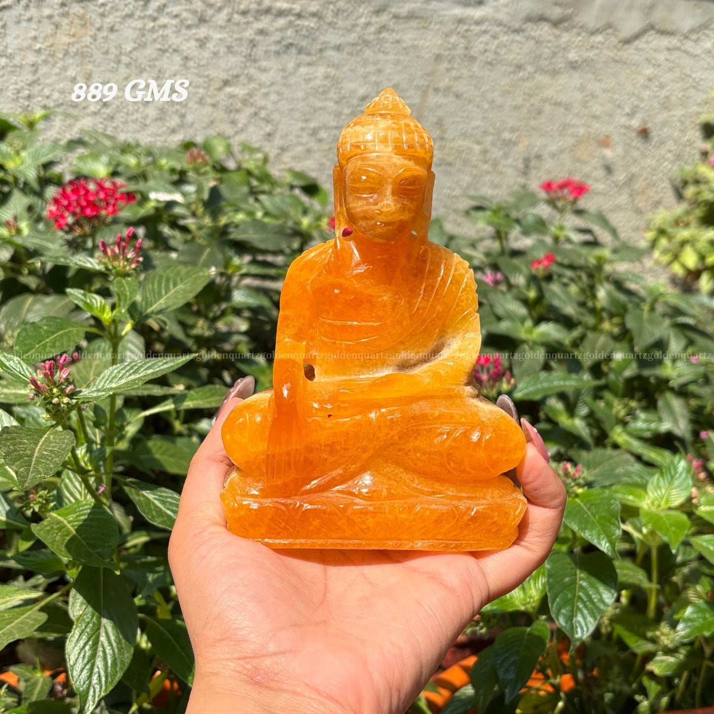 Hand holding a yellow calcite crystal Buddha statue with plants in the background Golden Quartz The Crystal Hub