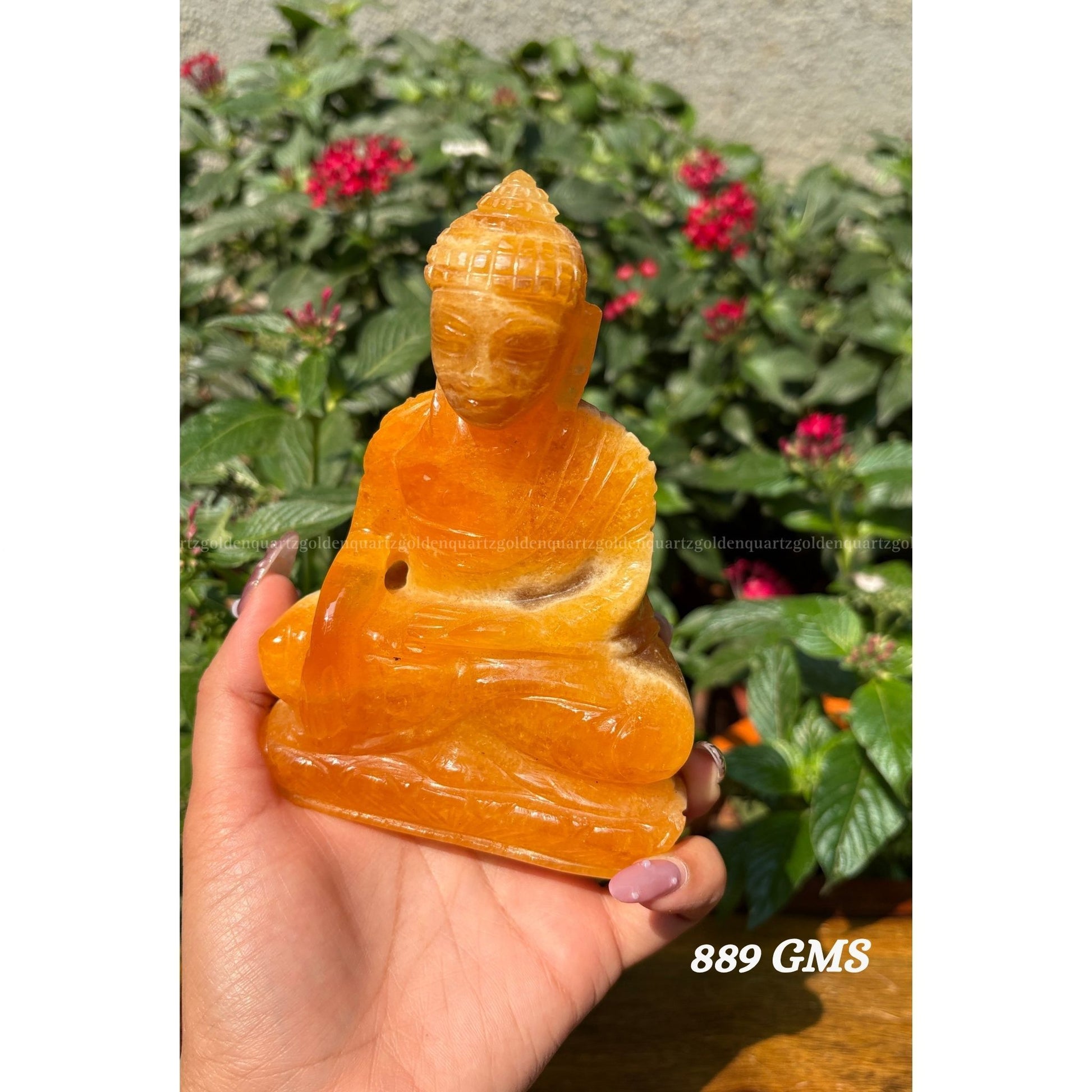 Hand holding a yellow calcite crystal Buddha statue with green plants and red flowers in the background Golden Quartz The Crystal Hub