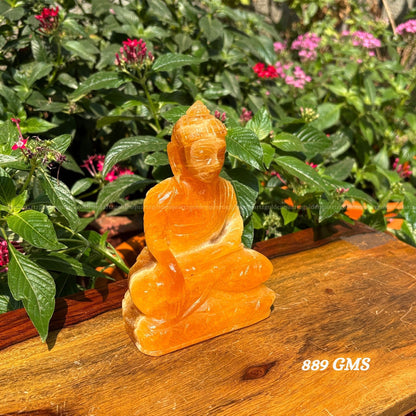 Orange crystal Buddha statue on a wooden surface with greenery in the background- Golden Quartz