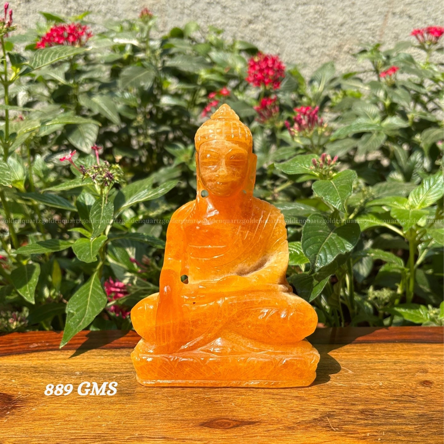 Orange stone Buddha statue on a wooden surface with greenery and flowers in the background